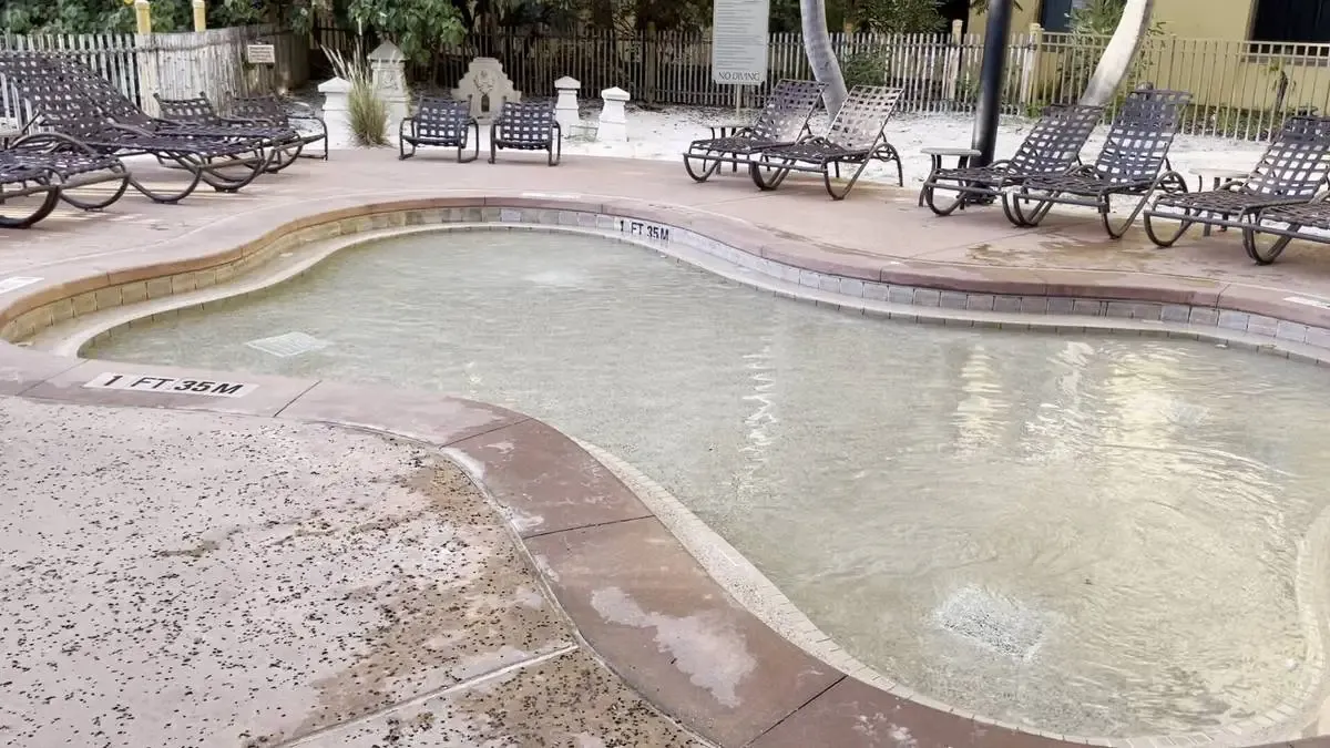 A shallow, curvy pool with a depth marked as 1 foot is surrounded by a wet deck and several brown lounge chairs. The area is bordered by a fence and some greenery, creating a relaxed outdoor setting.