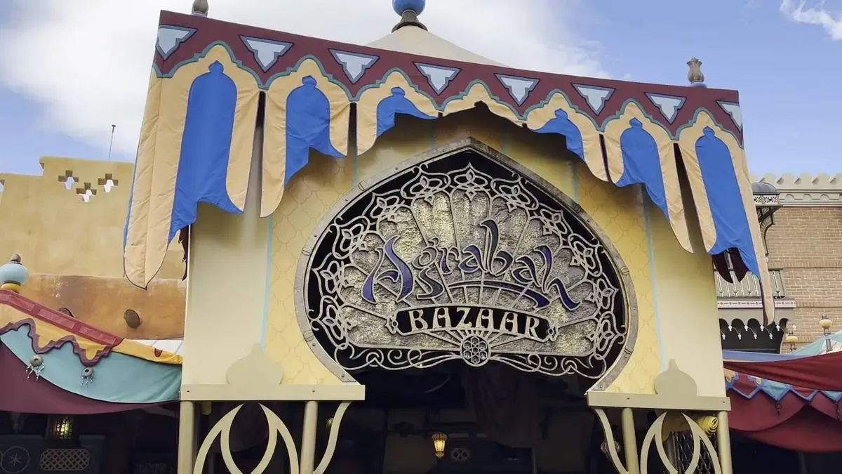 The image shows the entrance to the Agrabah Bazaar, featuring an ornate sign with intricate designs. The structure is adorned with decorative blue and beige fabric accents, giving it an exotic and inviting appearance.
