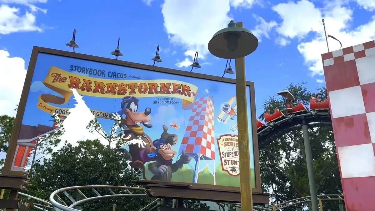 A sign for "The Barnstormer" ride at Storybook Circus features cartoon characters in a playful aviation theme. The roller coaster track curves nearby with red and white cars visible, set against a bright blue sky with fluffy clouds.