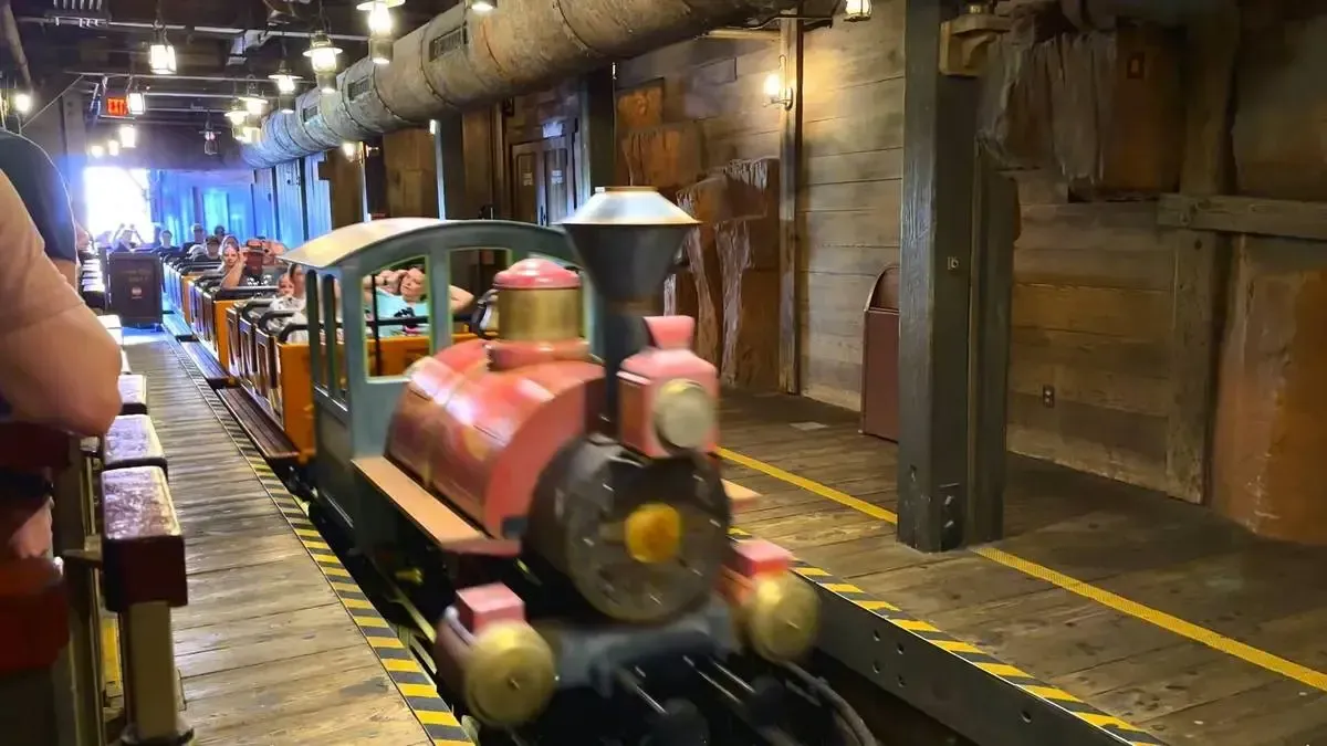 A colorful train ride with passengers is moving through a wooden-themed indoor station, featuring rustic walls and overhead pipes, creating a lively amusement park atmosphere.
