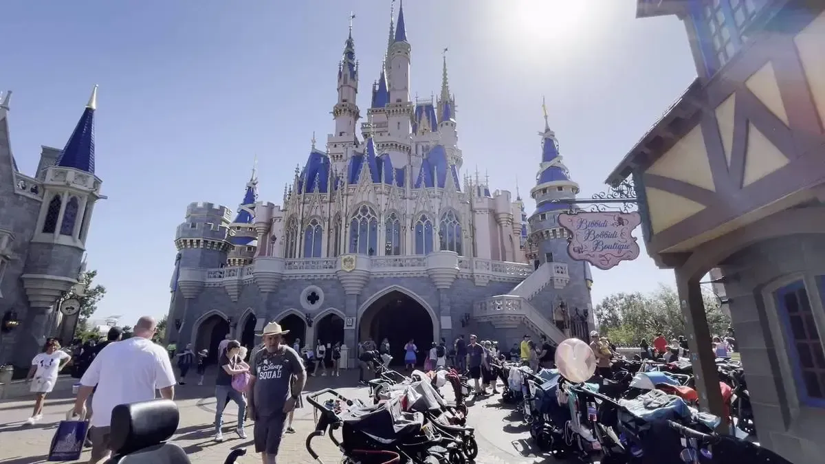 People stroll in front of Magic Kingdom castle with blue spires under a bright sky, surrounded by parked strollers and a sign for a boutique.