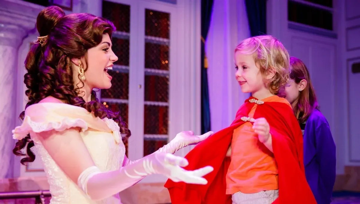 A woman dressed as a princess Belle interacts joyfully with a young child wearing a red cape and orange shirt. They are in a warmly lit room with ornate decor and large windows in the background.