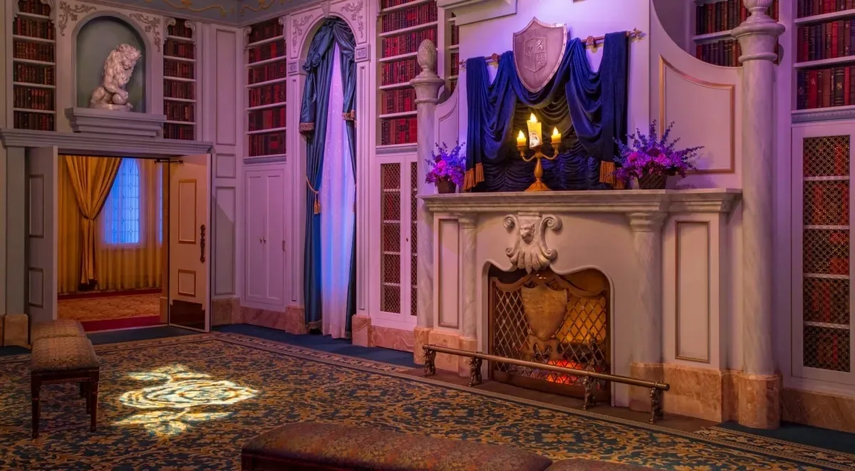 A richly decorated room features a grand fireplace adorned with blue drapery and a candelabra, next to purple flowers. Bookshelves filled with books surround the fireplace, and a statue of a lion perches above a doorway.