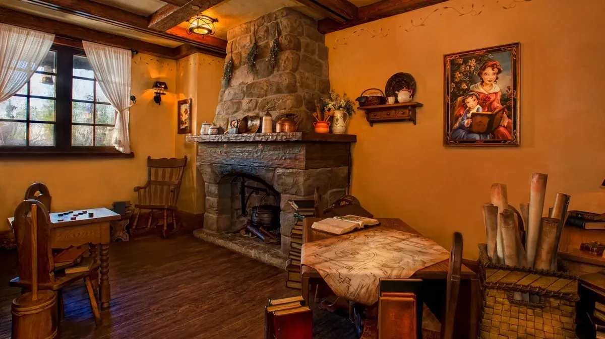 A cozy room features a rustic stone fireplace adorned with copper pots and a small shelf with decorative items. Wooden furniture, including a rocking chair and a table with an open book and map, creates a warm atmosphere.