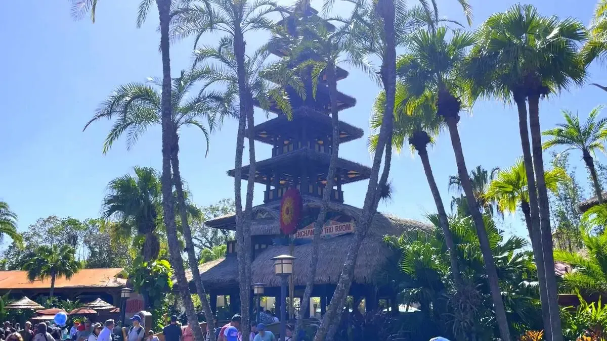 A tropical-themed building with a multi-tiered roof surrounded by tall palm trees under a clear blue sky. A crowd of people is gathered in front, enjoying the sunny day.