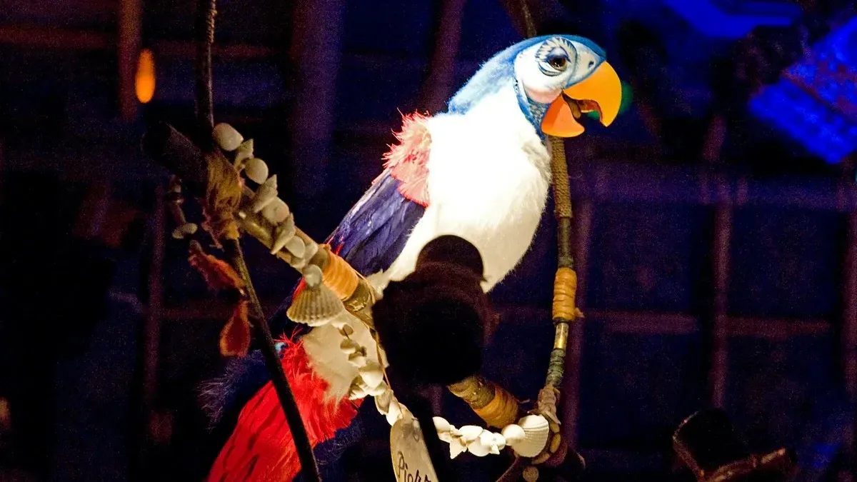 A colorful animatronic parrot with a vibrant orange beak and blue, white, and red feathers is perched on a decorated branch, set against a dimly lit background.