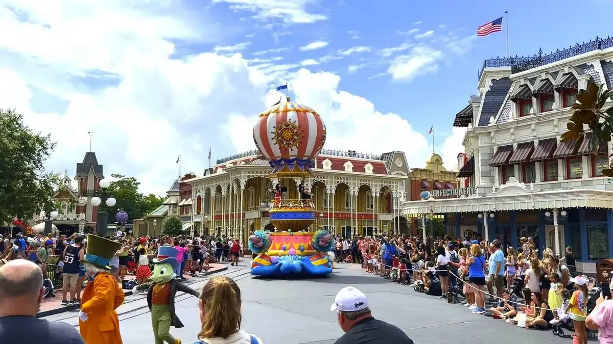 A colorful parade at a theme park features a vibrant float with a hot air balloon design, surrounded by costumed characters and enthusiastic spectators lining the street. The background showcases ornate buildings and a clear blue sky, creating a lively and festive atmosphere.