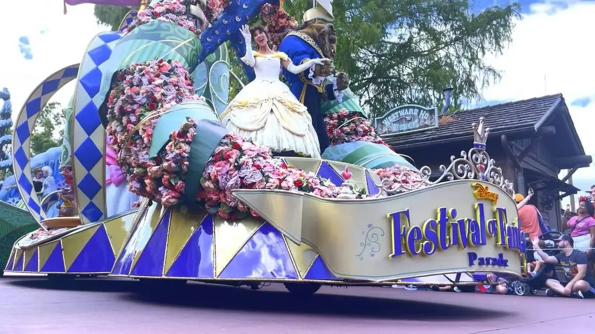 Costumed characters from "Snow White and the Seven Dwarfs," including the dwarfs and Snow White, parade down a theme park street lined with spectators. The scene is colorful and lively, with a whimsical float in the background and people capturing the moment on their phones.