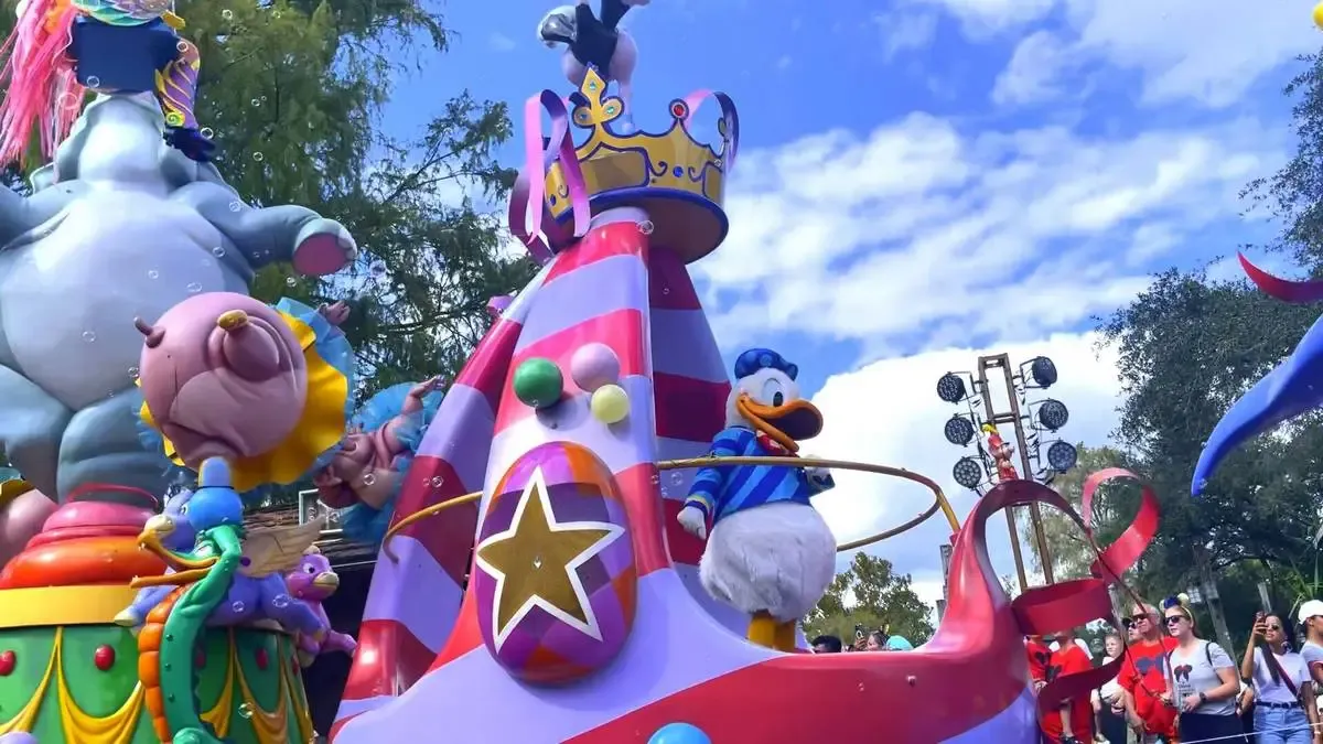 Costumed characters from "Snow White and the Seven Dwarfs," including the dwarfs and Snow White, parade down a theme park street lined with spectators. The scene is colorful and lively, with a whimsical float in the background and people capturing the moment on their phones.