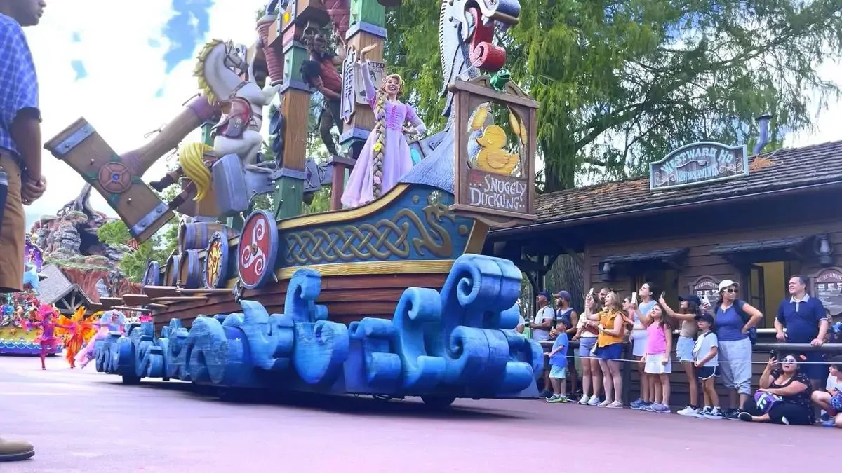 A colorful parade float with a whimsical boat design features a character in a purple dress waving to the crowd. The float, decorated with vibrant details and a "Snuggly Duckling" sign, passes by an excited audience on a theme park street.