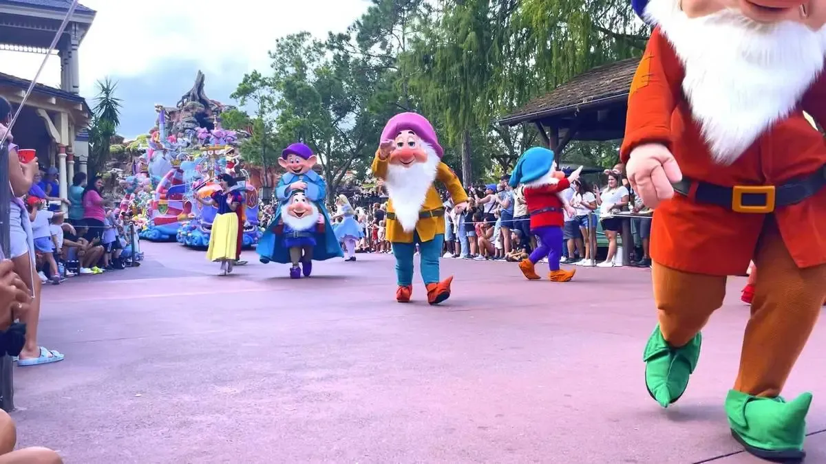 Costumed characters from "Snow White and the Seven Dwarfs," including the dwarfs and Snow White, parade down a theme park street lined with spectators. The scene is colorful and lively, with a whimsical float in the background and people capturing the moment on their phones.