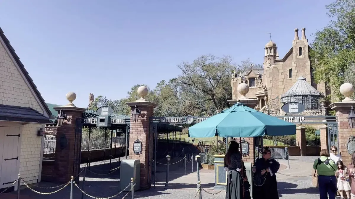 The image shows the entrance to a theme park attraction with a grand, mansion-like building in the background. The area is marked by brick pillars and ornate gates, with a teal umbrella providing shade for staff members. A few visitors are seen walking nearby, enjoying a clear, sunny day.