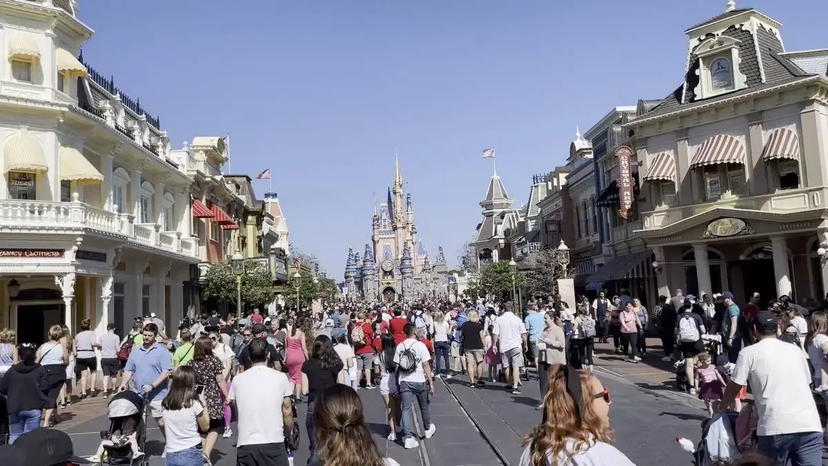 A bustling crowd walks along Main Street, USA, at a Disney theme park, with the iconic Cinderella Castle visible in the background. The street is lined with charming, vintage-style buildings under a clear blue sky.