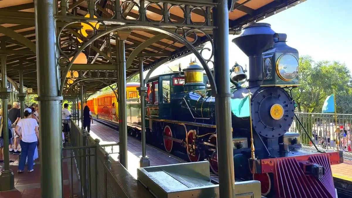 A vintage steam locomotive with a dark blue engine and red accents is parked at a station with ornate green metalwork. Passengers are waiting on the platform, and the train's yellow and orange passenger cars are visible in the background under a sunny sky.