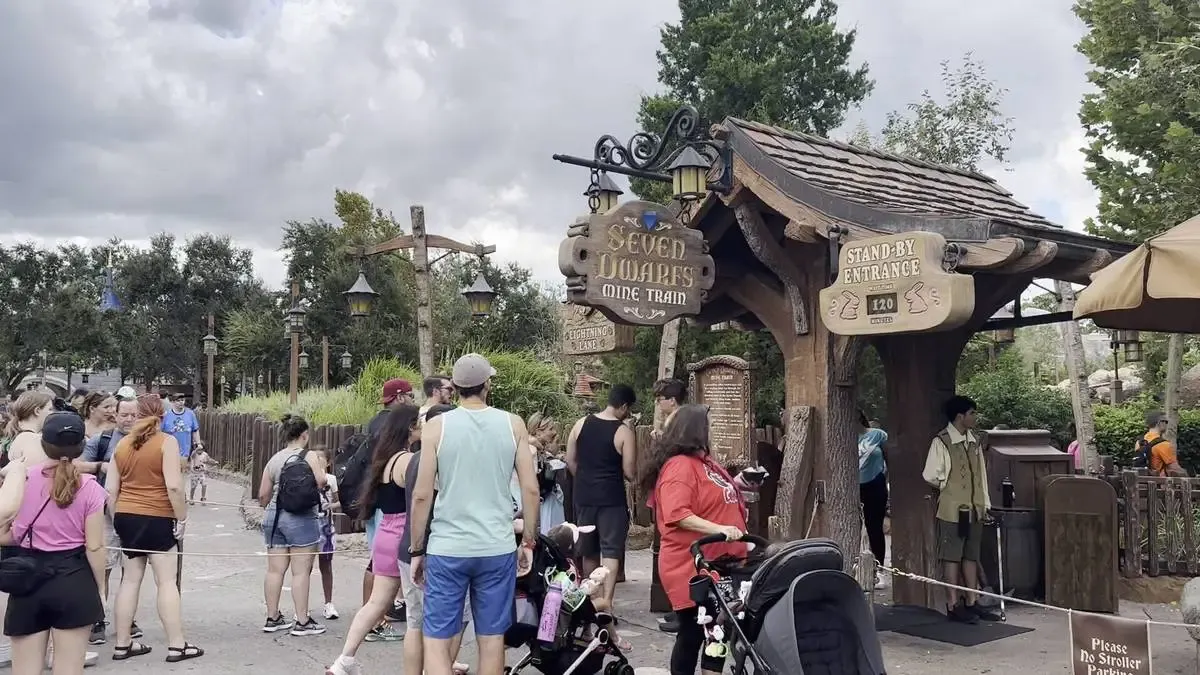 A crowd of people waits in line at the entrance of the Seven Dwarfs Mine Train ride, with a sign indicating a 120-minute standby wait time. The setting features rustic wooden structures and lush greenery under a cloudy sky.