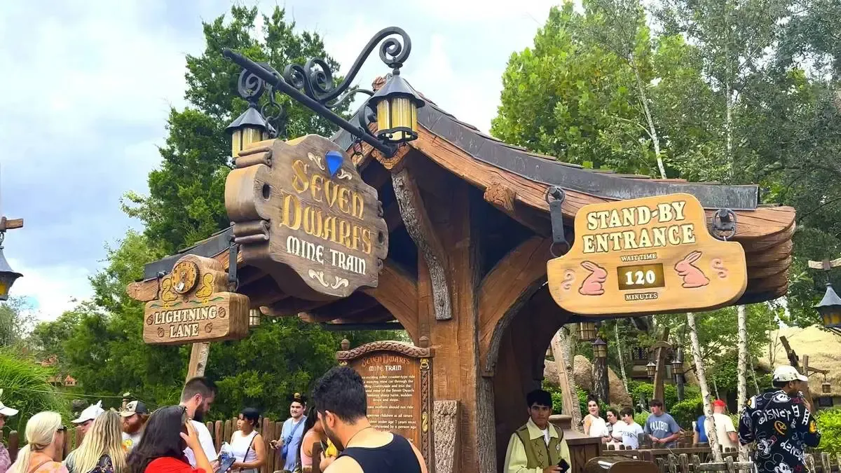 Visitors gather at the entrance of the Seven Dwarfs Mine Train ride, featuring rustic wooden indicating wait time for the ride, against a backdrop of lush greenery and cloudy skies.