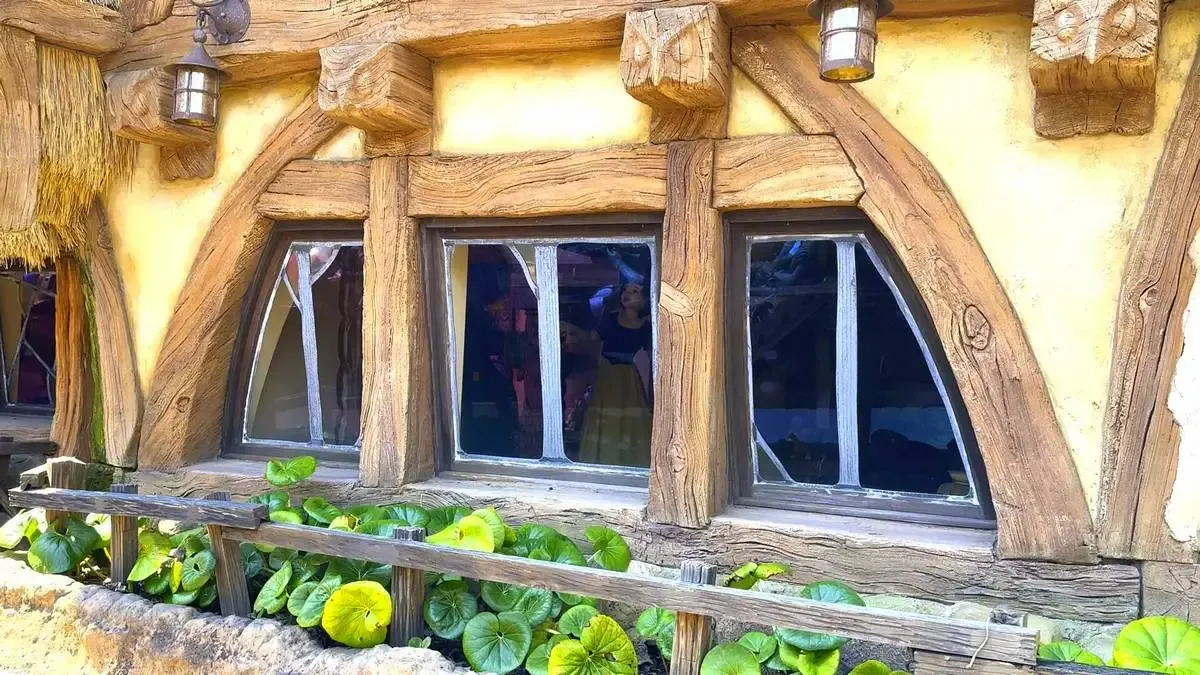 A charming, rustic cottage facade features three windows on a wooden frame with a slightly warped design. The walls are textured in a warm yellow with wooden beams creating a cozy, storybook aesthetic. Lush green plants line the base, and lanterns hang above.