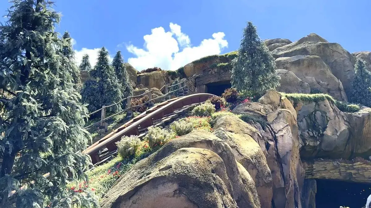 A wooden roller coaster track winds through a rocky landscape adorned with lush greenery and colorful flowers, surrounded by tall, evergreen trees under a bright blue sky with scattered clouds.
