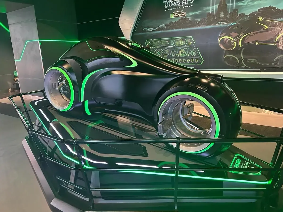 A futuristic black motorcycle with glowing green accents is displayed on a platform, surrounded by a black railing. The design features transparent wheels and is set against a backdrop of a digital-themed poster.