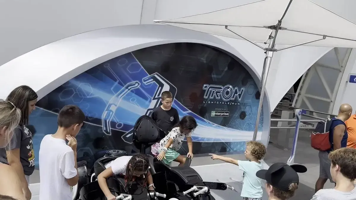 A group of children and adults gather around a TRON Lightcycle display, with some kids interacting with the exhibit. The background features a futuristic TRON-themed graphic. A white canopy provides shade over the area.