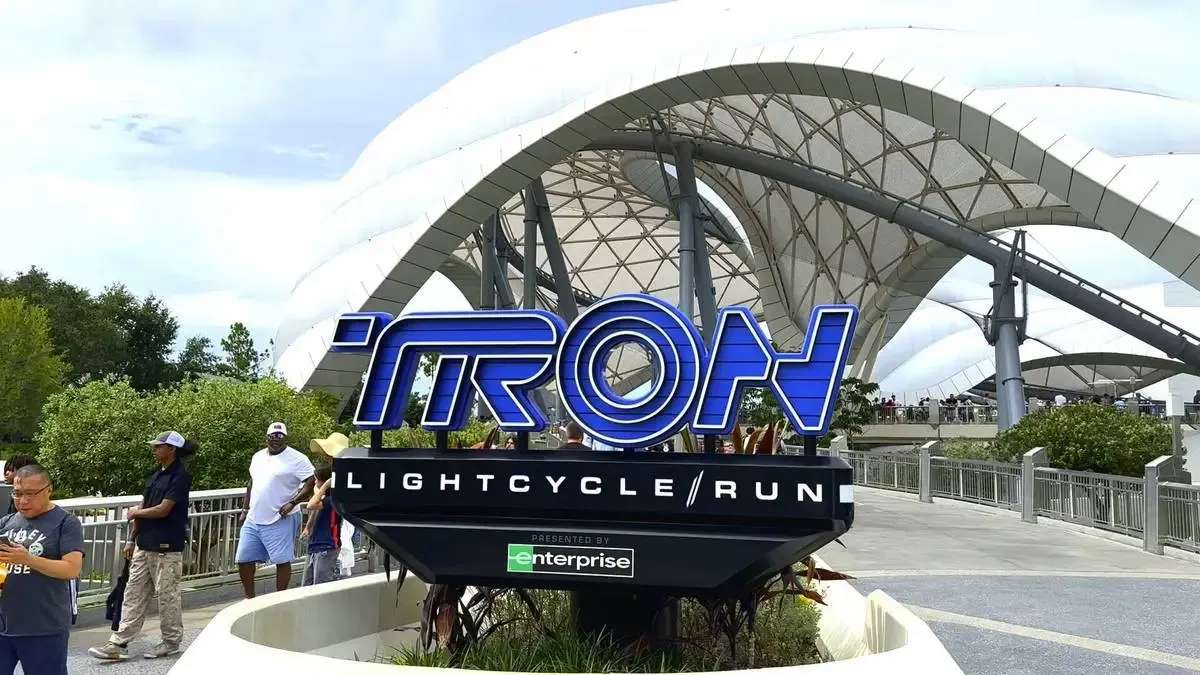 The image shows the entrance sign for "TRON Lightcycle / Run" at a Magic Kingdom, with a futuristic design featuring bold blue lettering. The background includes a sleek white structure and visitors walking nearby, set against a backdrop of greenery and a clear sky.