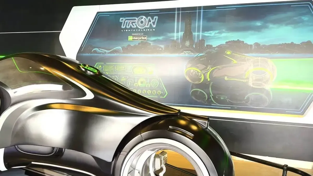 A sleek, futuristic motorcycle with a glossy black and silver finish is displayed in front of a digital screen featuring the "TRON Lightcycle Run" logo and a neon-lit cityscape.