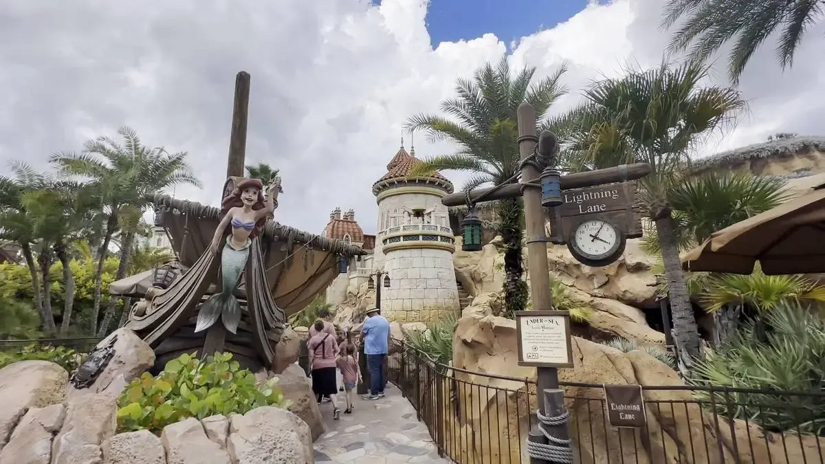 A statue of Ariel from the Little Mermaid is perched on a ship's bow in a themed amusement park area, surrounded by palm trees and rocky landscaping.