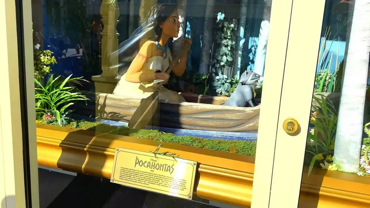 A display case features a detailed scene with Pocahontas in a canoe, accompanied by a raccoon. The background includes lush greenery and trees, creating a vibrant, natural setting. A plaque labeled "Pocahontas" is visible at the bottom.