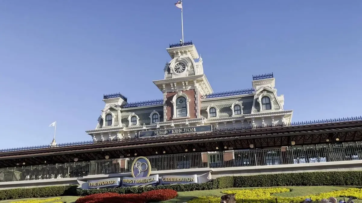 The iconic entrance of Magic Kingdom features a grand, Victorian-style train station with a clock tower, set against a clear blue sky. A banner celebrating "The World's Most Magical Celebration" is prominently displayed in front, surrounded by vibrant landscaping.