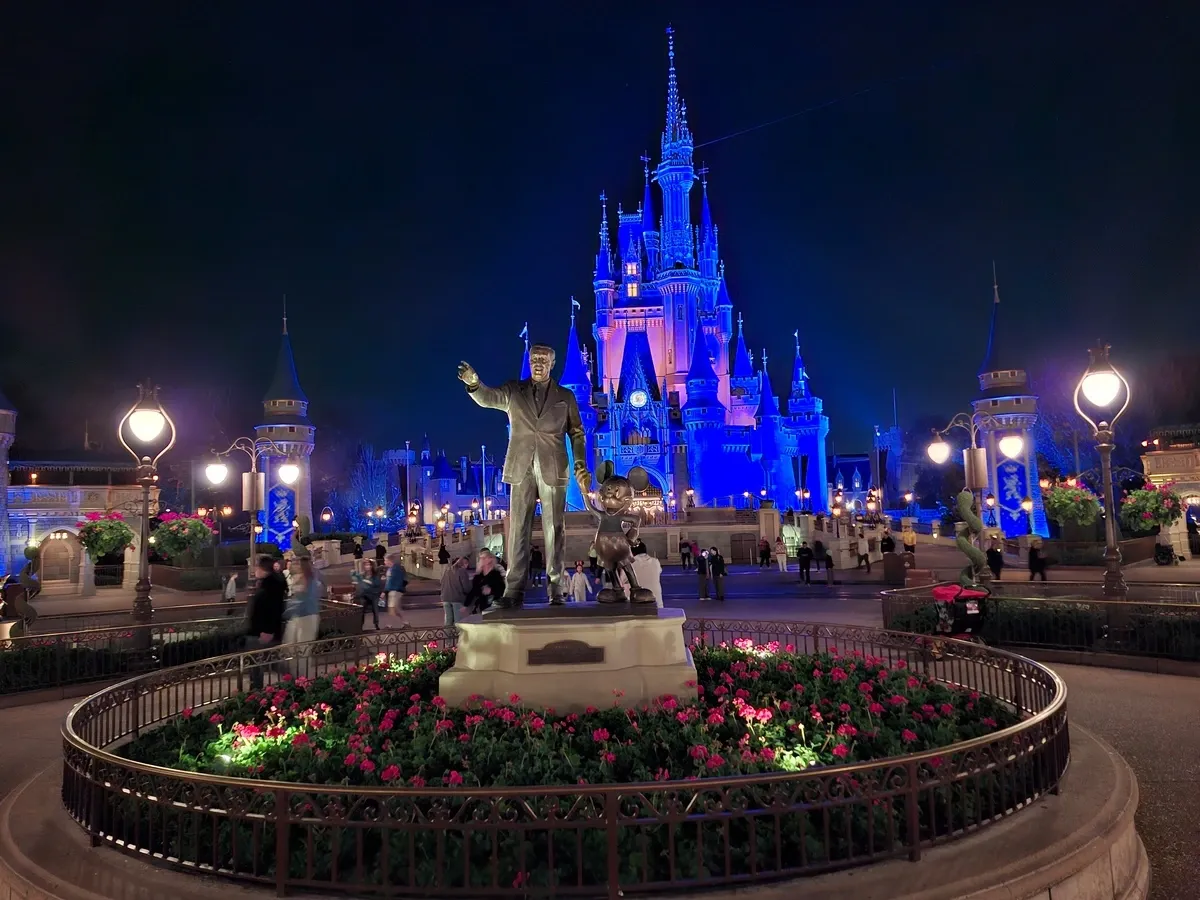 A nighttime scene at a theme park features a statue of a Walt Disney and Mickey Mouse standing together, surrounded by pink flowers. In the background, Magic Kingdom castle is illuminated in blue, with visitors strolling around the area.