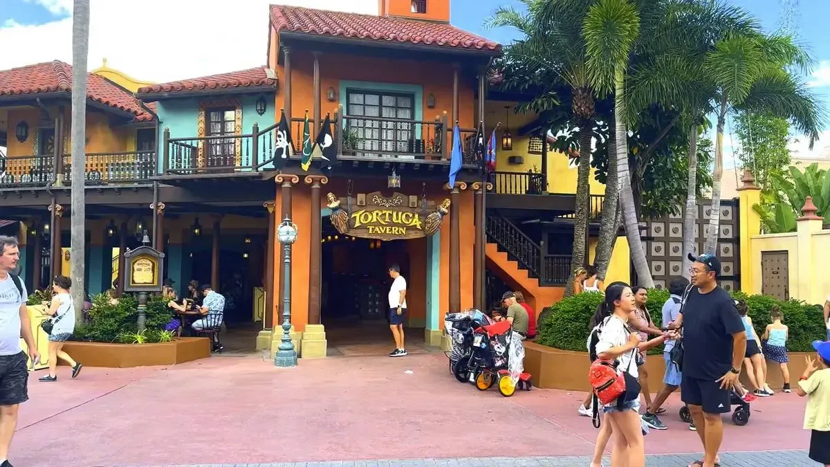 People stroll and relax outside the colorful Tortuga Tavern, featuring vibrant orange and blue walls, wooden balconies, and a red-tiled roof, with palm trees adding to the lively atmosphere.