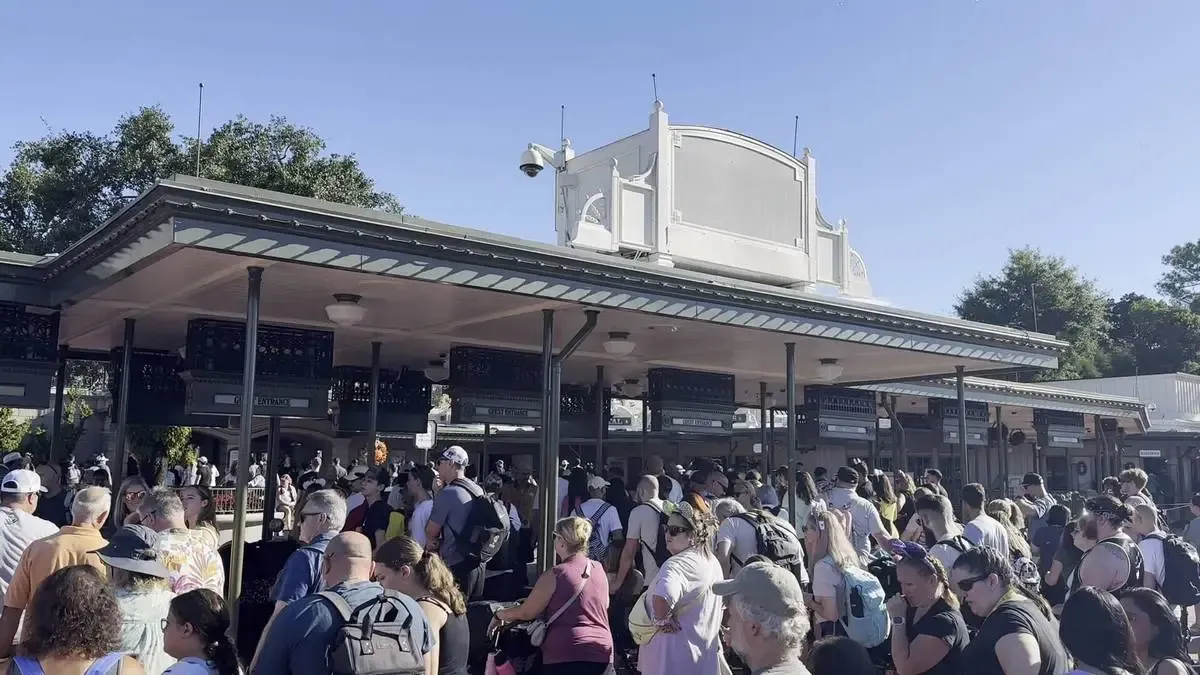 A large crowd of people gathers at the entrance of a theme park, waiting in line under a covered area with multiple ticket booths. 