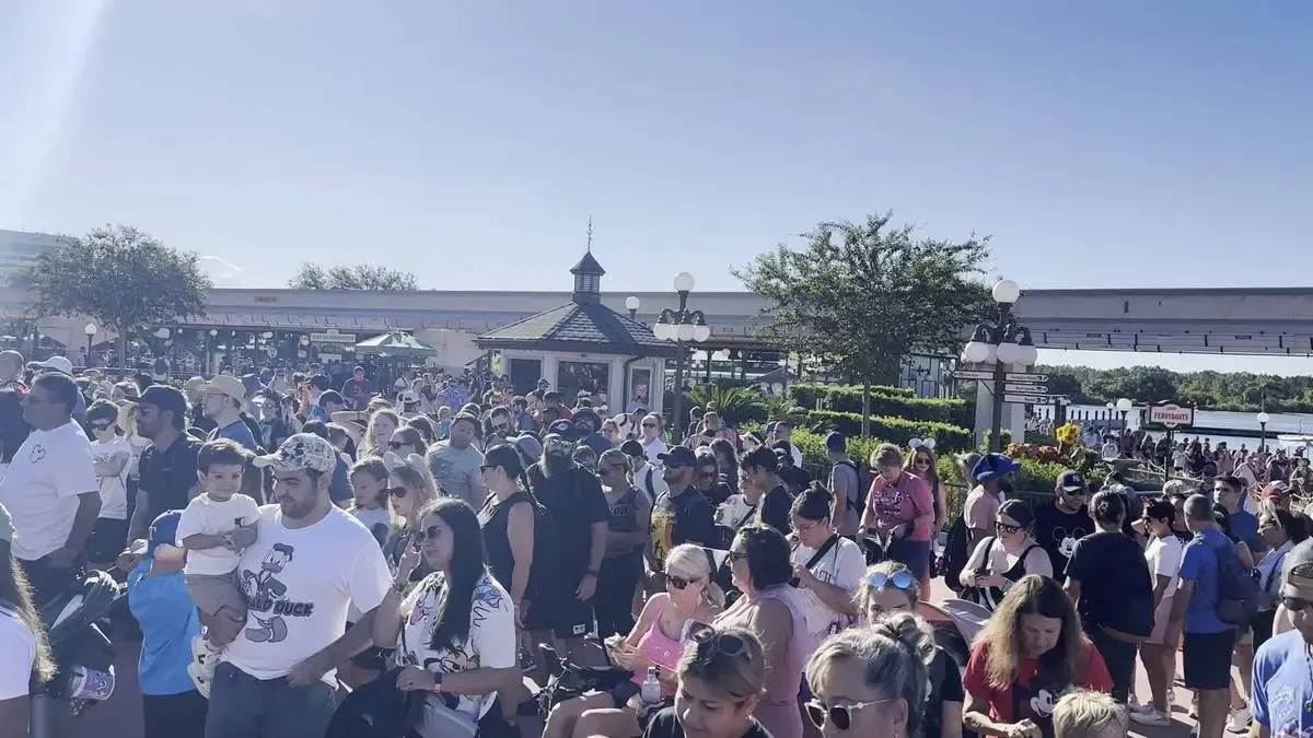 A large crowd of people gathered on a sunny day on the entrance of Magic Kingdom. In the background, a monorail track and a small pavilion are visible, along with trees and lampposts enhancing the lively atmosphere.
