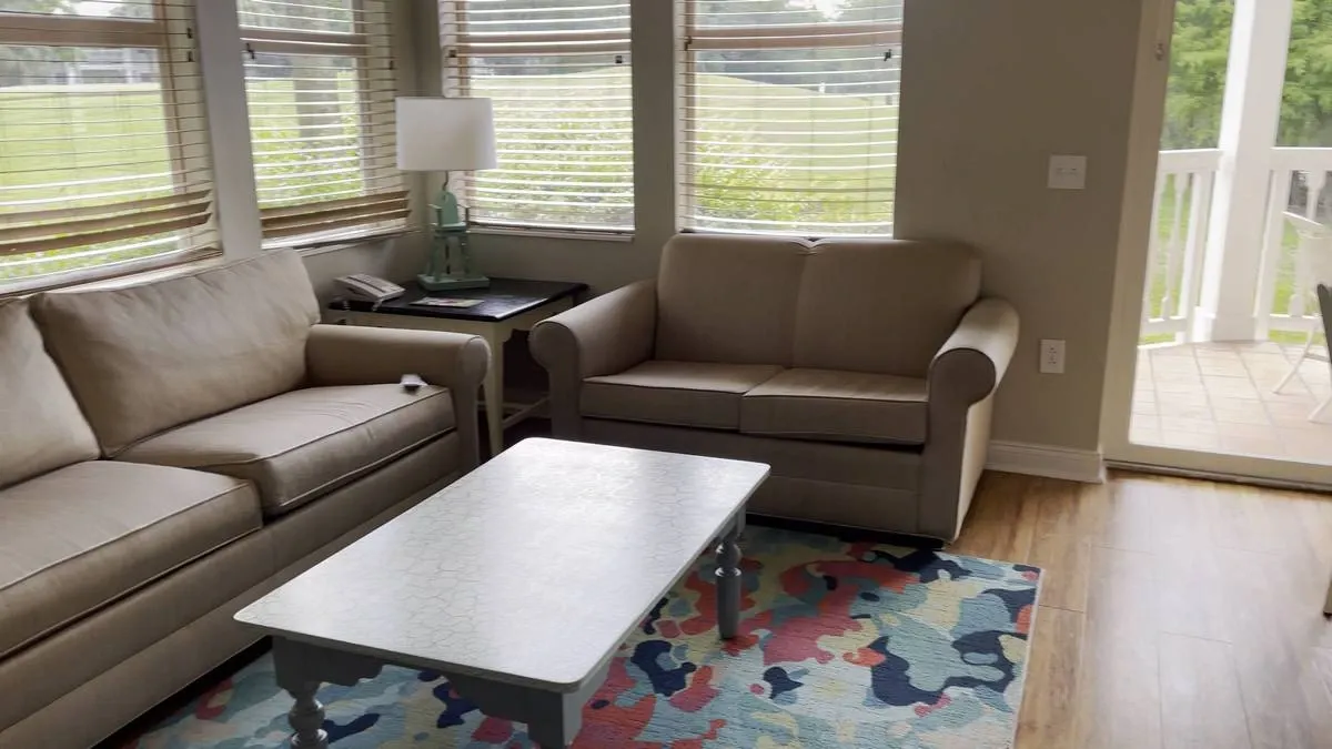A cozy living room features a beige sofa and loveseat, a white coffee table, and a colorful abstract rug. Large windows with wooden blinds allow natural light to fill the space, and a lamp sits on a side table.