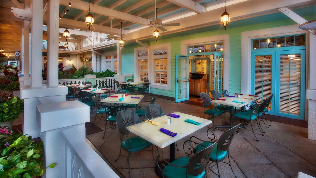 A charming outdoor restaurant patio features several tables with turquoise chairs and colorful napkins. The setting is enhanced by soft hanging lanterns and a pastel green and blue color scheme on the building.