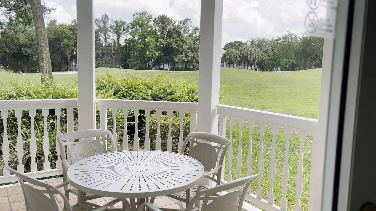A cozy patio features a round white table with four matching chairs, set against a backdrop of lush green grass and trees, framed by a white railing.