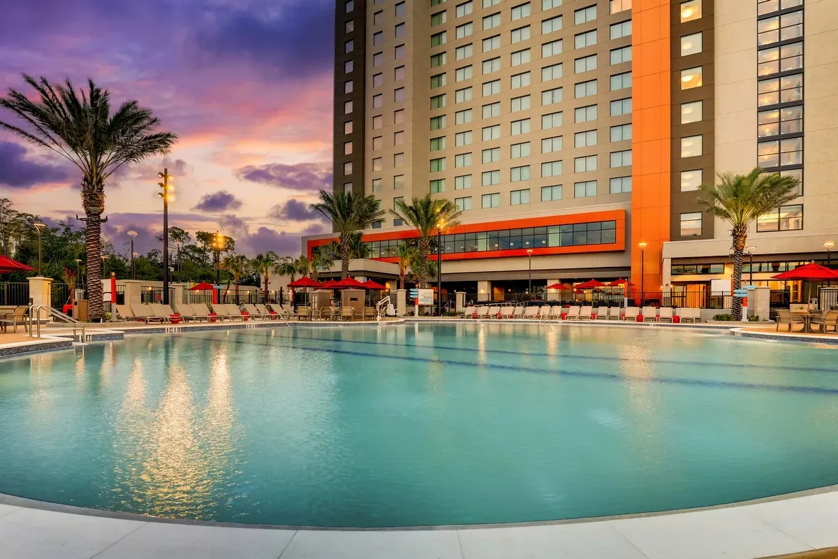 A large outdoor swimming pool is surrounded by lounge chairs and red umbrellas, set against the backdrop of a tall hotel building. Palm trees line the area, and the sky is a vibrant mix of purple and orange hues, suggesting a picturesque sunset.