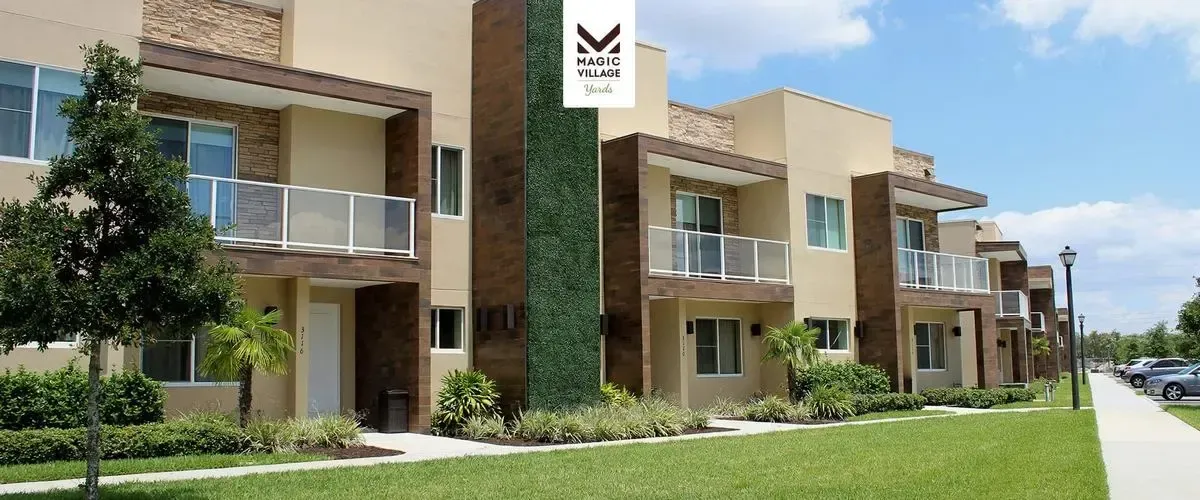 Modern townhouses at Magic Village Yards feature a sleek design with beige and brown exteriors, glass balconies, and lush landscaping, set against a clear blue sky.