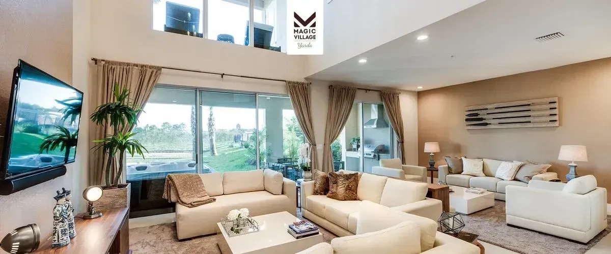 A spacious living room features cream color sofas, a large TV mounted on the wall, and a glass coffee table. The room is lit with natural light from the large sliding glass doors leading to a patio with a view of greenery. 