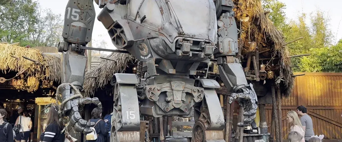 A large, detailed robot statue stands prominently in an outdoor setting, surrounded by people. The robot features intricate mechanical parts and is marked with the number "15" on its arms. Behind it, there are rustic structures with thatched roofs, and trees in the background.