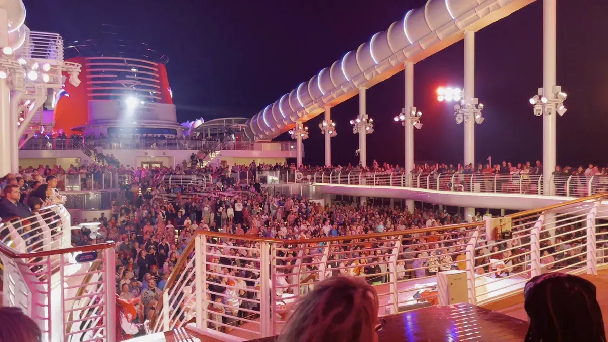 A large crowd gathers on the deck of a cruise ship at night, illuminated by bright lights and surrounded by white railings. The ship's distinctive red funnel is visible in the background, adding to the lively atmosphere.