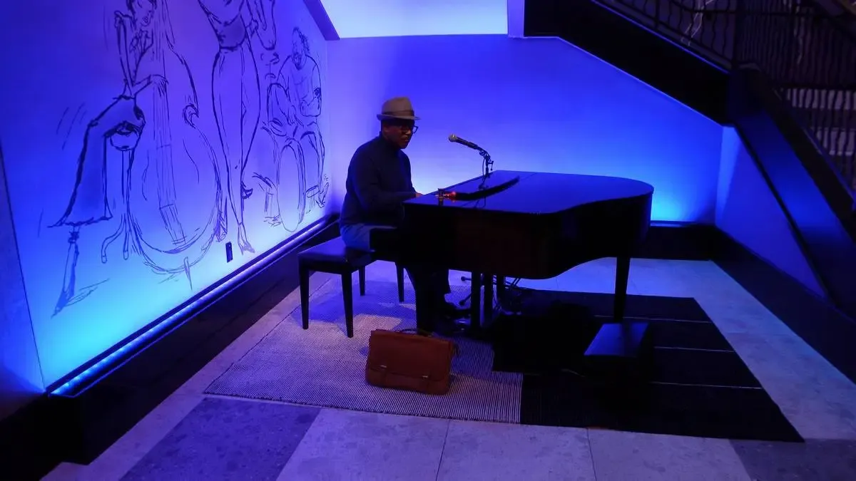 A musician wearing a hat plays a grand piano under soft blue lighting, with a microphone set up nearby. The background features a mural of jazz musicians, enhancing the artistic atmosphere. A brown briefcase rests on the floor beside the piano.