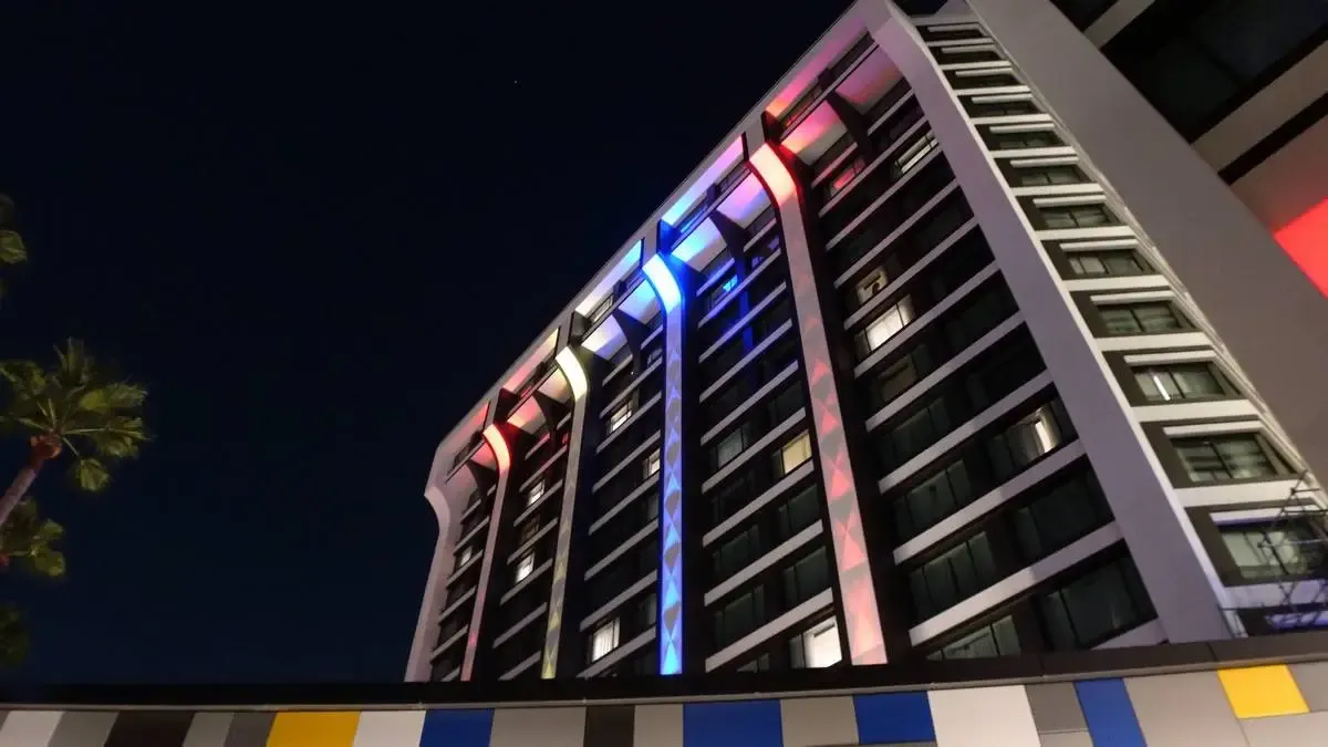 A modern hotel building illuminated at night with vibrant red and blue lights accentuating its vertical lines. The structure features numerous windows, and a palm tree is visible against the dark sky, adding a touch of greenery to the scene.