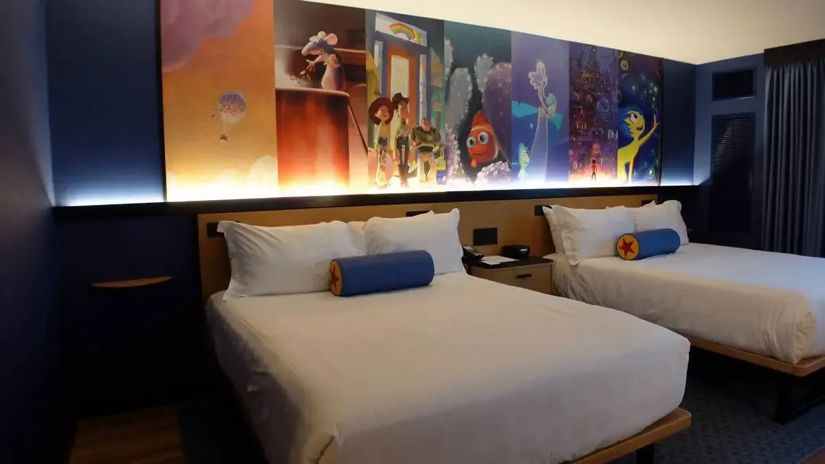 A hotel room featuring two beds with white linens, each adorned with a blue cylindrical pillow featuring a yellow star. Above the beds, a vibrant mural showcases various animated characters in a colorful, illuminated display.