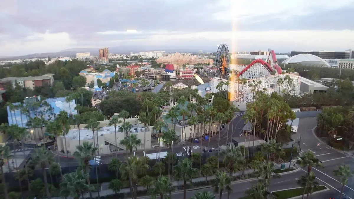 Aerial view of Disneyland park featuring a large roller coaster with red tracks and a Ferris wheel with a sunburst design. The park is surrounded by greenery and palm trees, with various colorful attractions visible in the background under a cloudy sky.