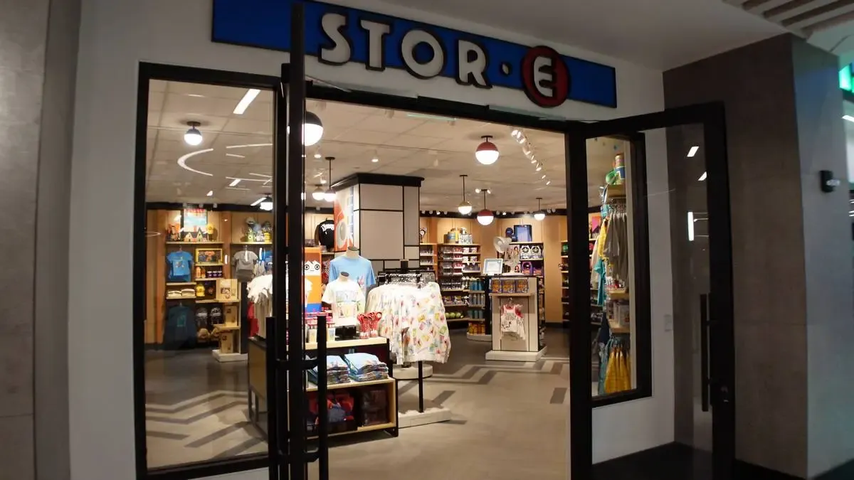 A store entrance with glass doors leads into an interior filled with colorful merchandise. T-shirts and other clothing items are displayed at the front, with shelves of various products visible in the background. The store sign above reads "STOR-E" in bold letters.