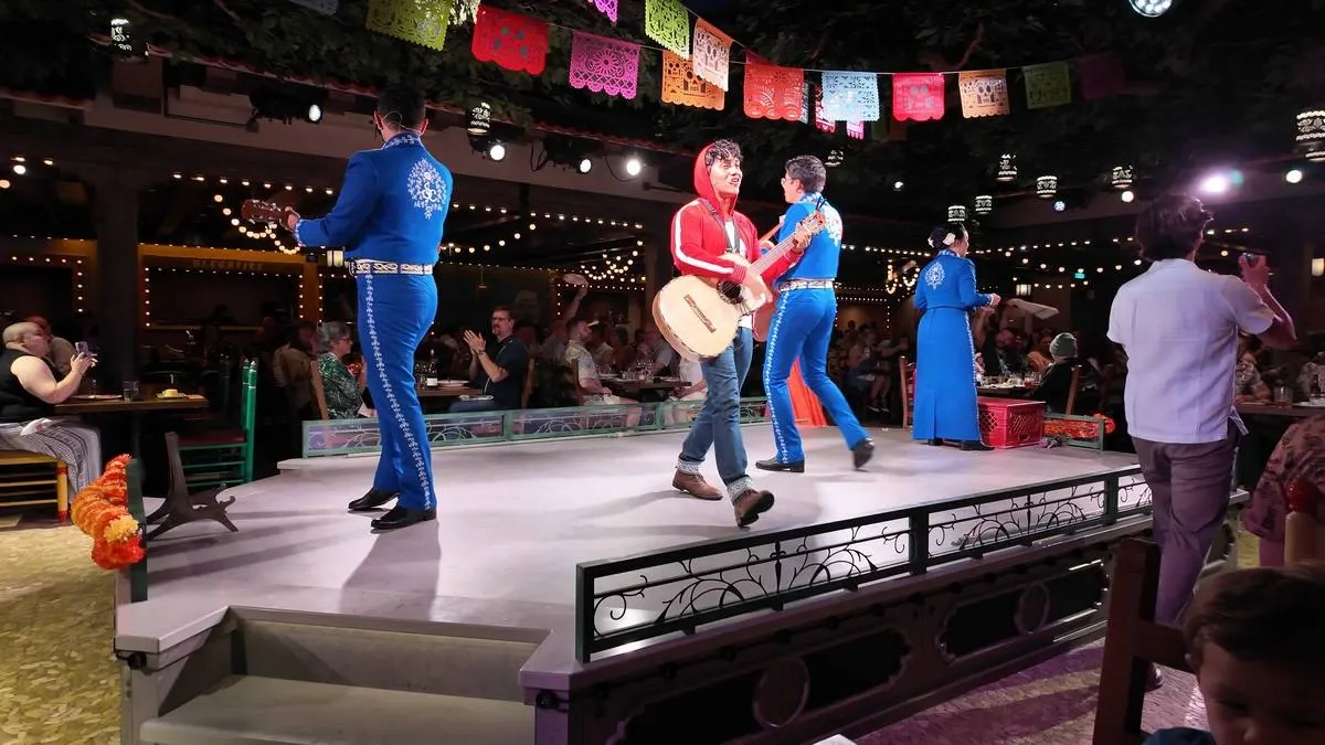 A lively stage performance features four musicians, three in bright blue mariachi outfits and one in a red hoodie, playing guitars. Colorful papel picado banners hang above, and the audience at surrounding tables enjoys the festive atmosphere under warm string lights.