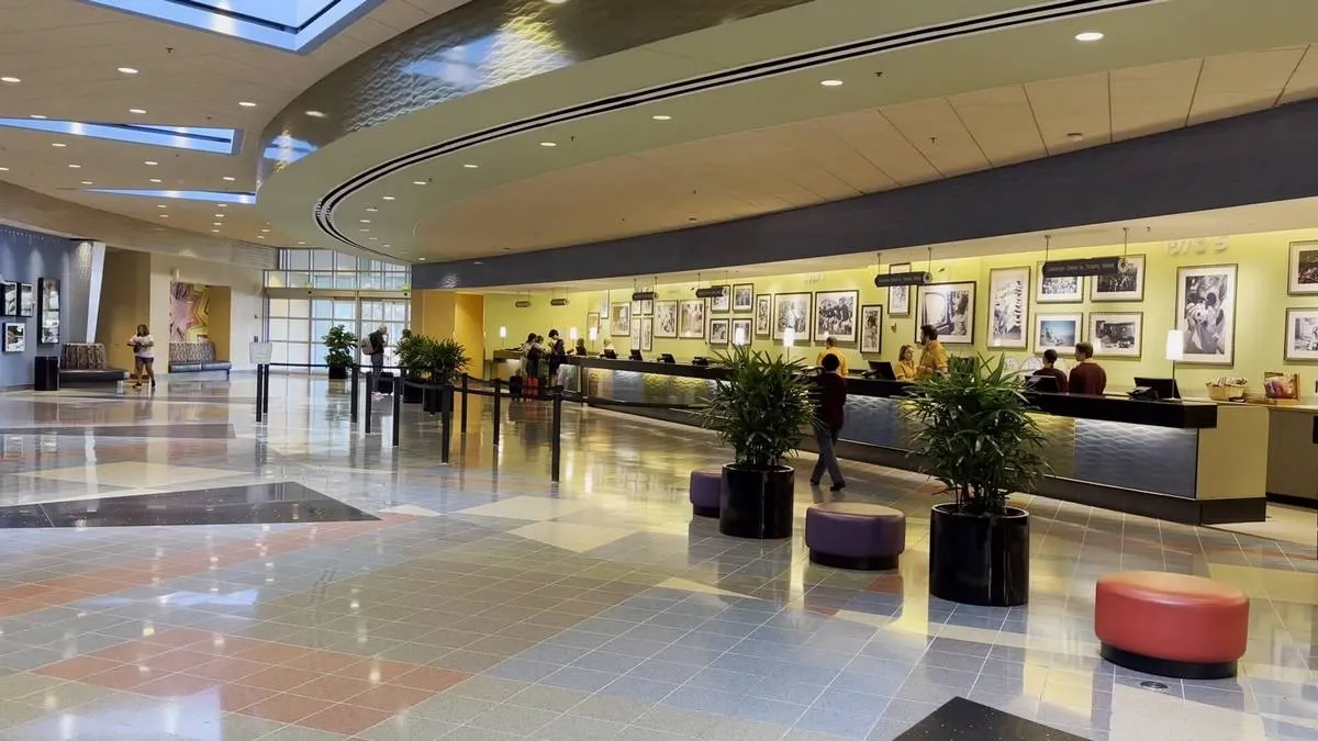 A spacious hotel lobby features a long reception desk with several people checking in. The floor has a colorful tile pattern, and the ceiling boasts skylights that let in natural light.