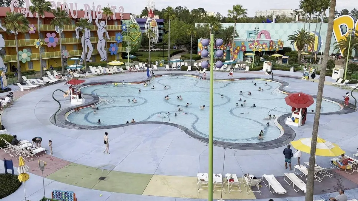 A hotel pool area features a large swimming pool in a flower shape with numerous people enjoying the water. Surrounding the pool are colorful buildings adorned with retro flower designs and palm trees.