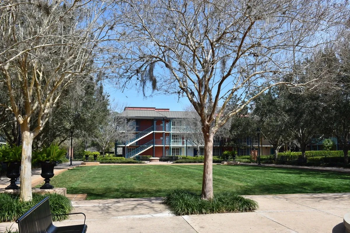 A peaceful courtyard features a grassy area bordered by leafless trees, with a red-brick building and blue staircases in the background. Benches and planters with green foliage line the paved walkways.